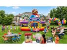 A large, candy-themed inflatable play center featuring giant lollipops, candy cane arches, and a gumball machine topper. The backyard scene shows children playing in the inflatable while families enjoy a party with popcorn, cotton candy, and snow cone sta