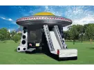 A wide, vibrant photograph of a colossal, silver and black alien spacecraft-themed inflatable bouncer set up on a massive field of green grass. The inflatable features a prominent yellow dome top, a line of flashing red "lights" around the rim, a large bl